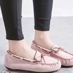 May include: A pair of light pink moccasin-style loafers with brown trim and a bow detail. The shoes are made of a soft material and have a stitched design around the toe. The image shows the shoes being worn with black pants.