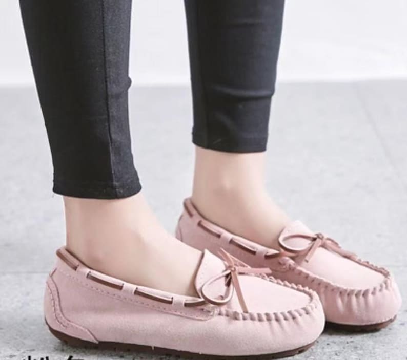 May include: A pair of light pink moccasin-style loafers with brown trim and a bow detail. The shoes are made of a soft material and have a stitched design around the toe. The image shows the shoes being worn with black pants.