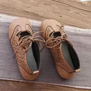May include: A pair of tan-colored, lace-up shoes with intricate cut-out detailing. The shoes have a rounded toe and a low heel. The laces are a matching tan color. The shoes are made of a soft material.