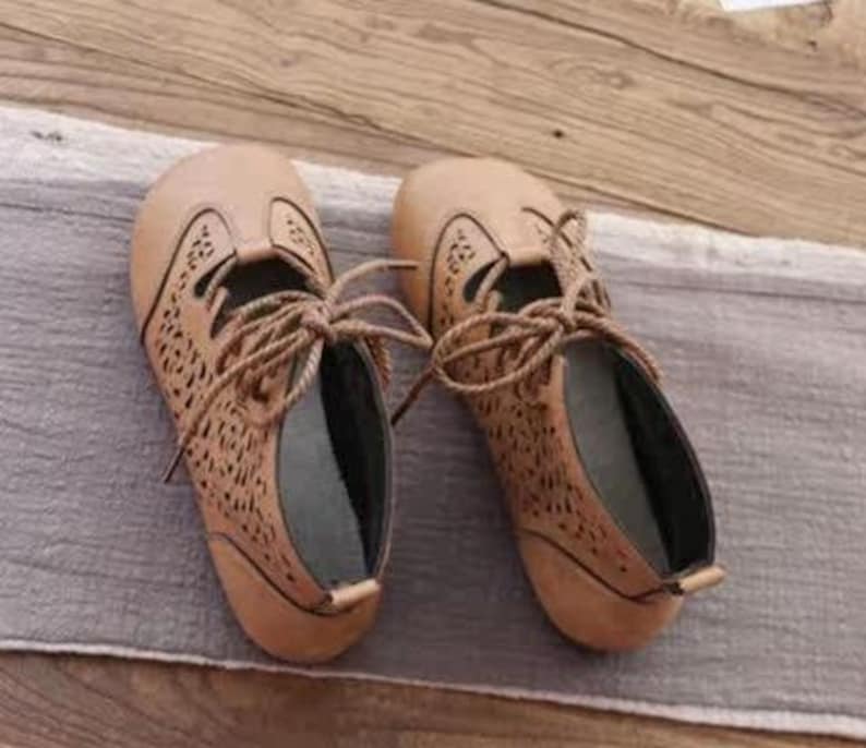 May include: A pair of tan-colored, lace-up shoes with intricate cut-out detailing. The shoes have a rounded toe and a low heel. The laces are a matching tan color. The shoes are made of a soft material.