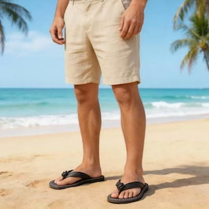Puede incluir: Pantalones cortos de lino color beige y chanclas de cuero negro con detalle floral. La persona est&aacute; de pie en una playa de arena con el oc&eacute;ano al fondo. Los pantalones cortos tienen bolsillos.