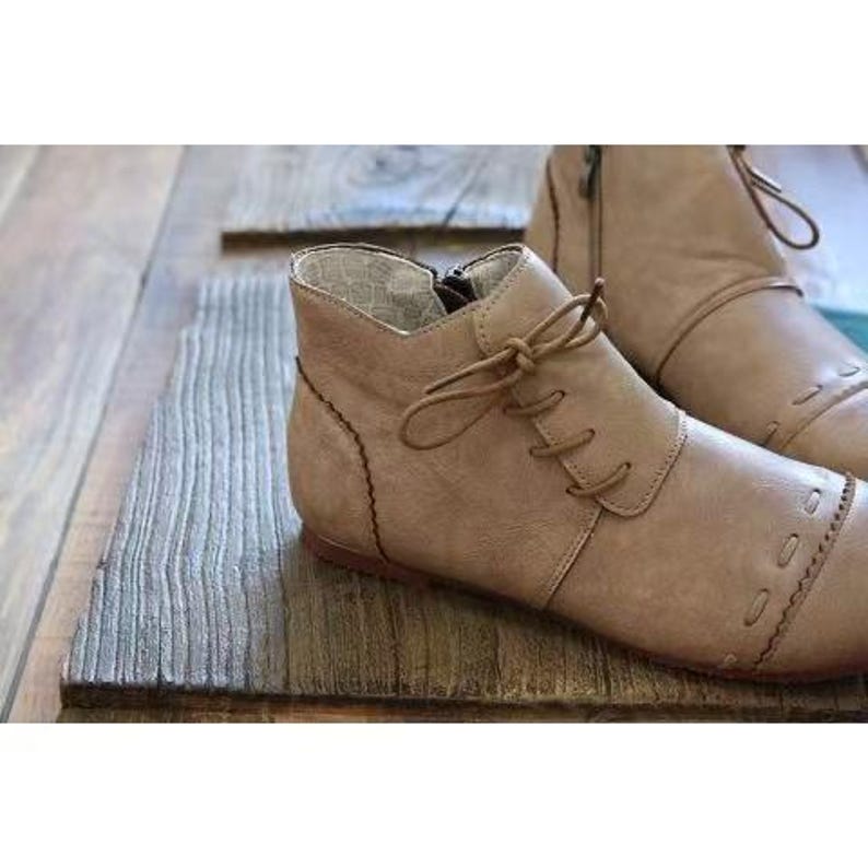 May include: Tan leather ankle boots with a lace-up design and decorative stitching. The boots have a side zipper and are set on a wooden surface. These boots are a stylish footwear option.
