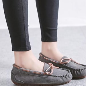 May include: Gray suede moccasin-style loafers with brown leather trim and a bow detail. The shoes have a rounded toe and a stitched design around the edge. The soles are a dark brown color.