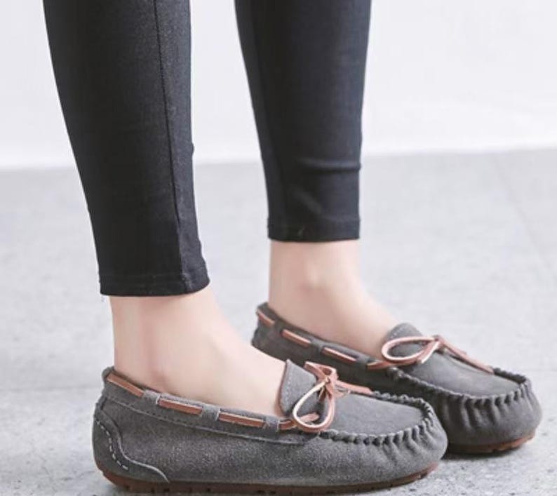 May include: Gray suede moccasin-style loafers with brown leather trim and a bow detail. The shoes have a rounded toe and a stitched design around the edge. The soles are a dark brown color.