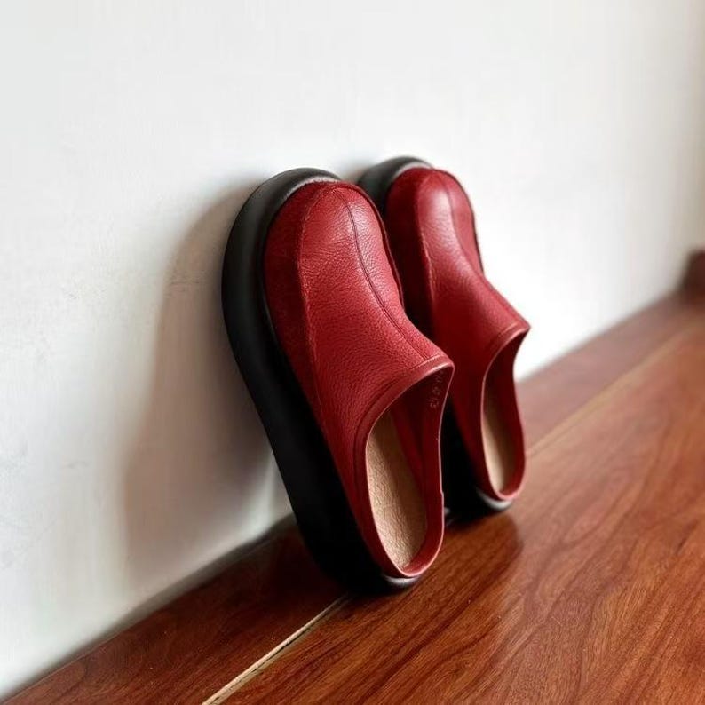 Puede incluir: Un par de zapatos sin cordones de cuero rojo con suela negra. Los zapatos est&aacute;n apoyados contra una pared blanca y est&aacute;n colocados sobre una superficie de madera. Los zapatos tienen punta redonda y tac&oacute;n bajo.
