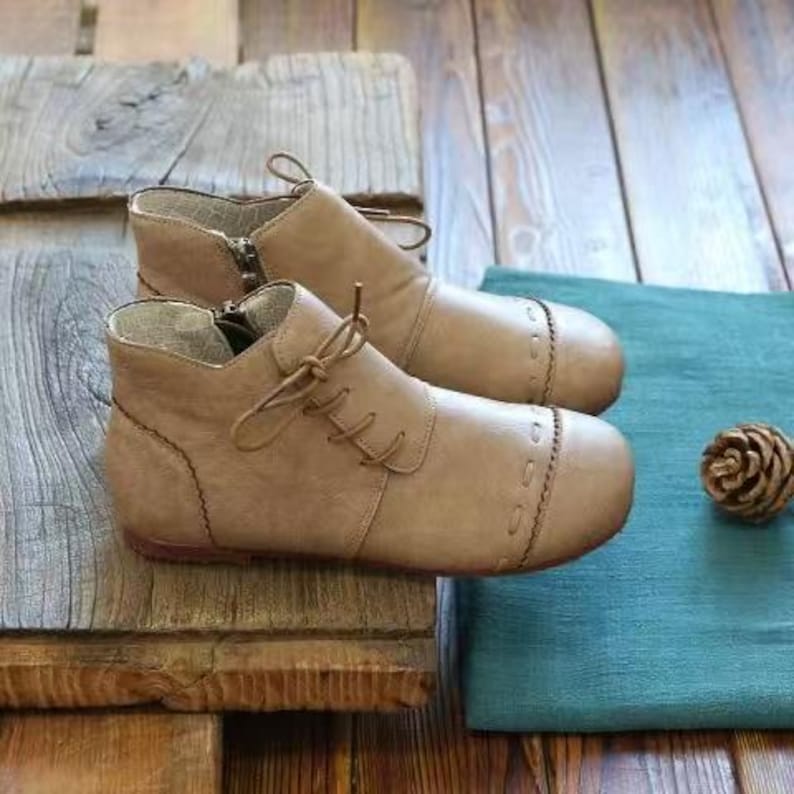 May include: A pair of tan leather ankle boots with laces and zippers. The boots have a rounded toe and decorative stitching. They are placed on a wooden surface next to a teal cloth and a pinecone.
