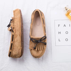 May include: Tan suede moccasin-style shoes with dark brown accents and fringe detailing. The shoes feature a rounded toe, a bow, and a stitched design around the sole. The soles are brown.
