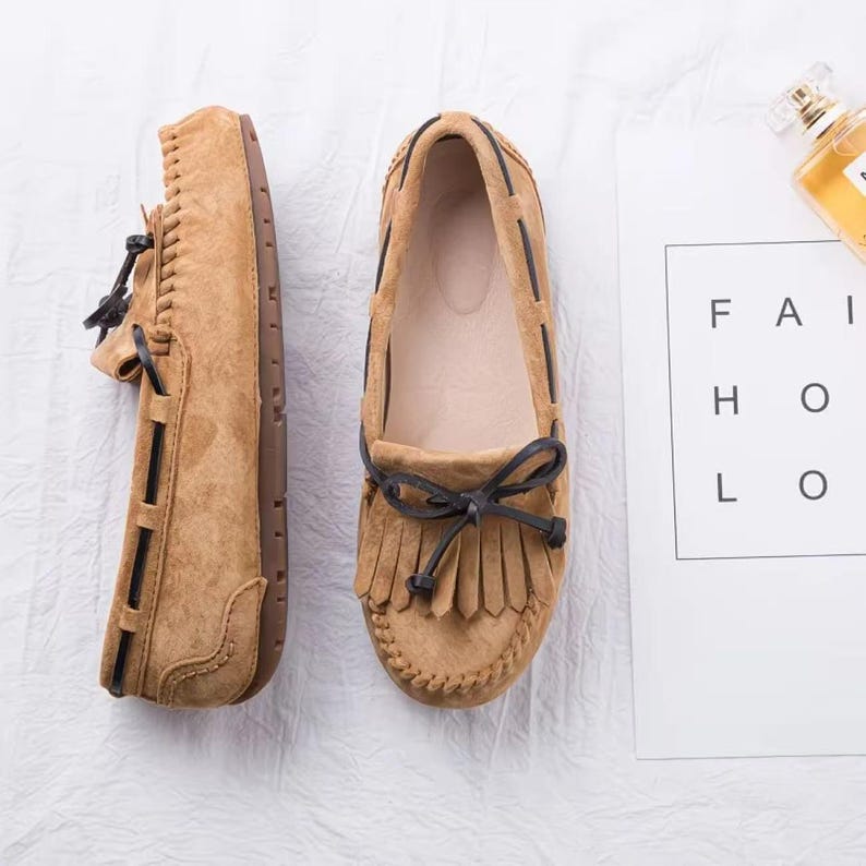 May include: Tan suede moccasin-style shoes with dark brown accents and fringe detailing. The shoes feature a rounded toe, a bow, and a stitched design around the sole. The soles are brown.