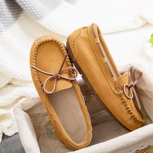 May include: A pair of mustard-colored suede moccasin-style loafers. The shoes feature brown laces tied in bows and are accented with decorative stitching. The soles are a dark brown color. The shoes are displayed in a woven basket.