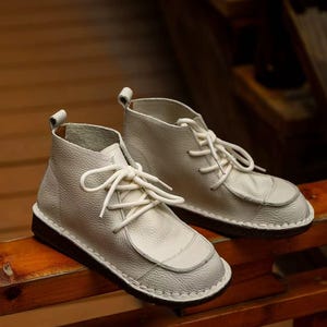 Pu&ograve; includere: Un paio di stivaletti alla caviglia in pelle bianca con lacci bianchi. Gli stivali hanno un design a punta arrotondata e una suola marrone scuro. Le scarpe sono su una superficie di legno.