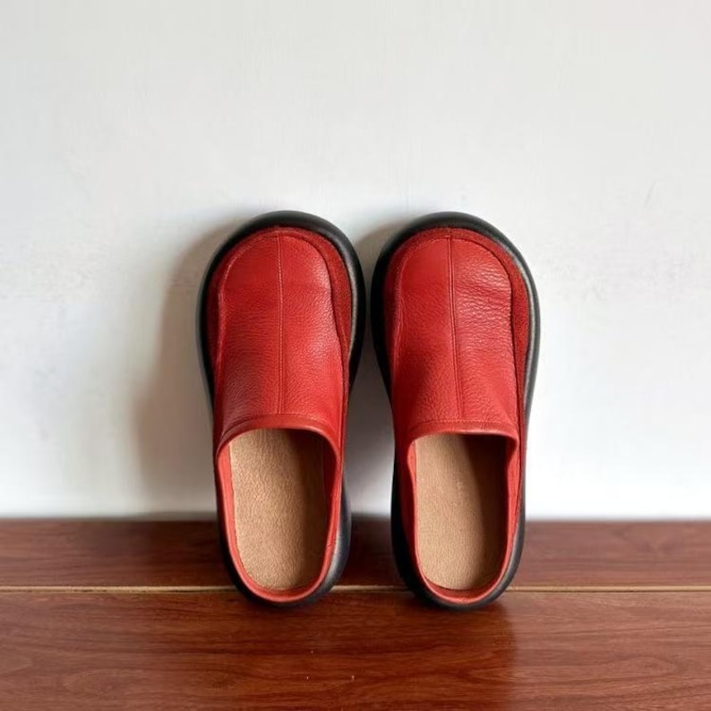 Puede incluir: Un par de zapatos sin cordones de cuero rojo con suela negra. Los zapatos tienen una puntera redondeada y una superficie lisa y texturizada. El interior es de color marr&oacute;n claro.