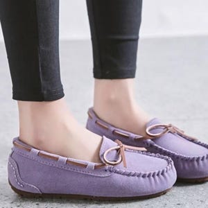 May include: A pair of lavender moccasin-style loafers with brown leather accents. The shoes have a rounded toe, a stitched sole, and a decorative bow. The shoes are worn with black pants.