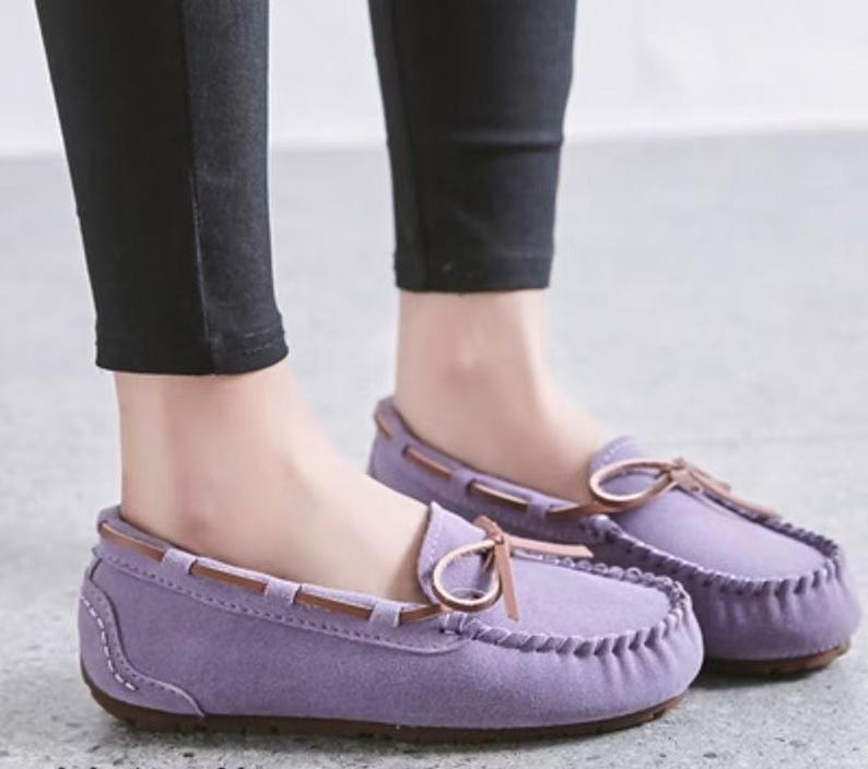 May include: A pair of lavender moccasin-style loafers with brown leather accents. The shoes have a rounded toe, a stitched sole, and a decorative bow. The shoes are worn with black pants.