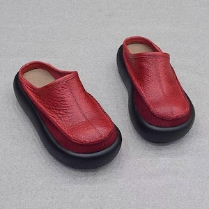 Könnte beinhalten: Ein Paar rote Lederclogs mit schwarzer Sohle. Die Clogs haben eine runde Spitze und ein Slip-on-Design. Das Obermaterial besteht aus strukturiertem rotem Leder, die Sohle ist dick und schwarz.