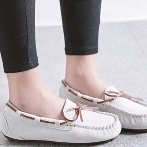 May include: White moccasin-style loafers with brown accents and a bow detail. The shoes have a stitched design around the edges and a brown sole. The loafers are made of a soft, suede-like material.