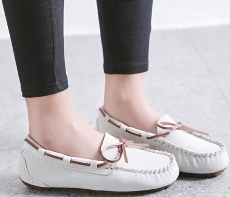 May include: White moccasin-style loafers with brown accents and a bow detail. The shoes have a stitched design around the edges and a brown sole. The loafers are made of a soft, suede-like material.