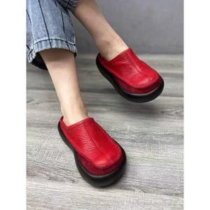 Puede incluir: Zuecos de cuero rojo con suela negra. Los zuecos tienen una puntera redondeada y una parte superior de cuero texturizado. Los zapatos se est&aacute;n usando, con los pies y la parte inferior de las piernas visibles. Los zuecos son un estilo llamativo.