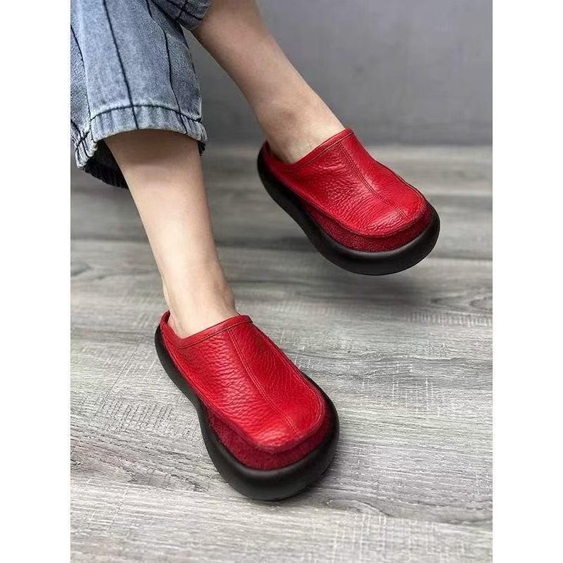Puede incluir: Zuecos de cuero rojo con suela negra. Los zuecos tienen una puntera redondeada y una parte superior de cuero texturizado. Los zapatos se est&aacute;n usando, con los pies y la parte inferior de las piernas visibles. Los zuecos son un estilo llamativo.