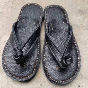 Puede incluir: Un par de chanclas de cuero negro con una superficie texturizada. Las sandalias presentan un dise&ntilde;o de correa de tanga con un detalle anudado y est&aacute;n cosidas con un hilo marr&oacute;n claro contrastante en los bordes.