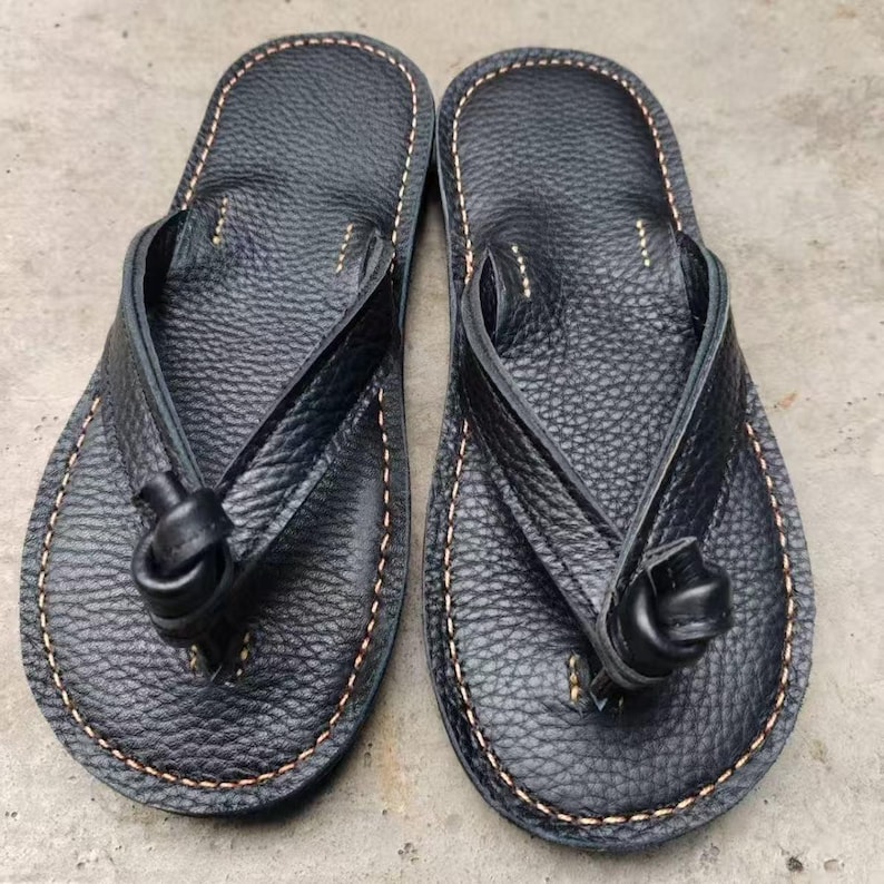 Puede incluir: Un par de chanclas de cuero negro con una superficie texturizada. Las sandalias presentan un dise&ntilde;o de correa de tanga con un detalle anudado y est&aacute;n cosidas con un hilo marr&oacute;n claro contrastante en los bordes.