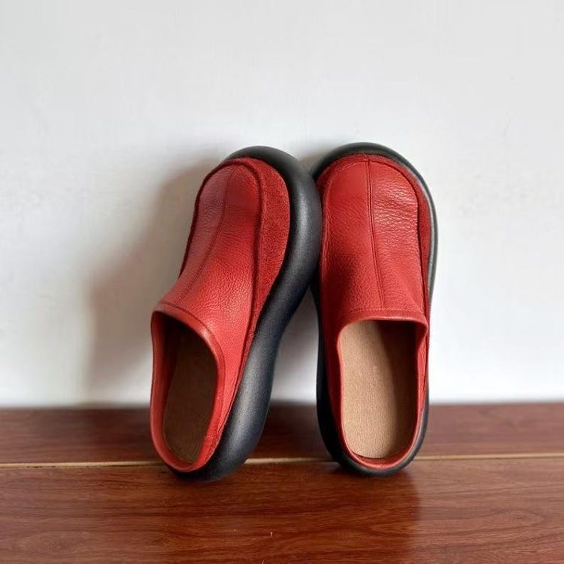 Puede incluir: Un par de zapatos sin cordones de cuero rojo con punta redondeada y suela negra gruesa. Los zapatos se exhiben sobre una superficie de madera sobre un fondo blanco. Dise&ntilde;o minimalista.