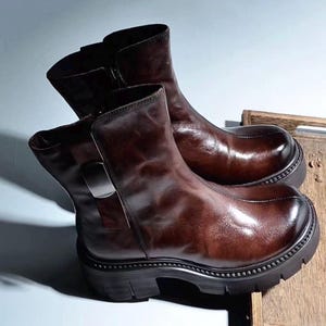 以下が含まれることがあります： サイドジッパーと分厚い黒いソールが付いた、茶色の革製アンクルブーツのペア。ブーツは光沢のある仕上がりで、丸いつま先です。ブーツは木製の表面に表示されています。