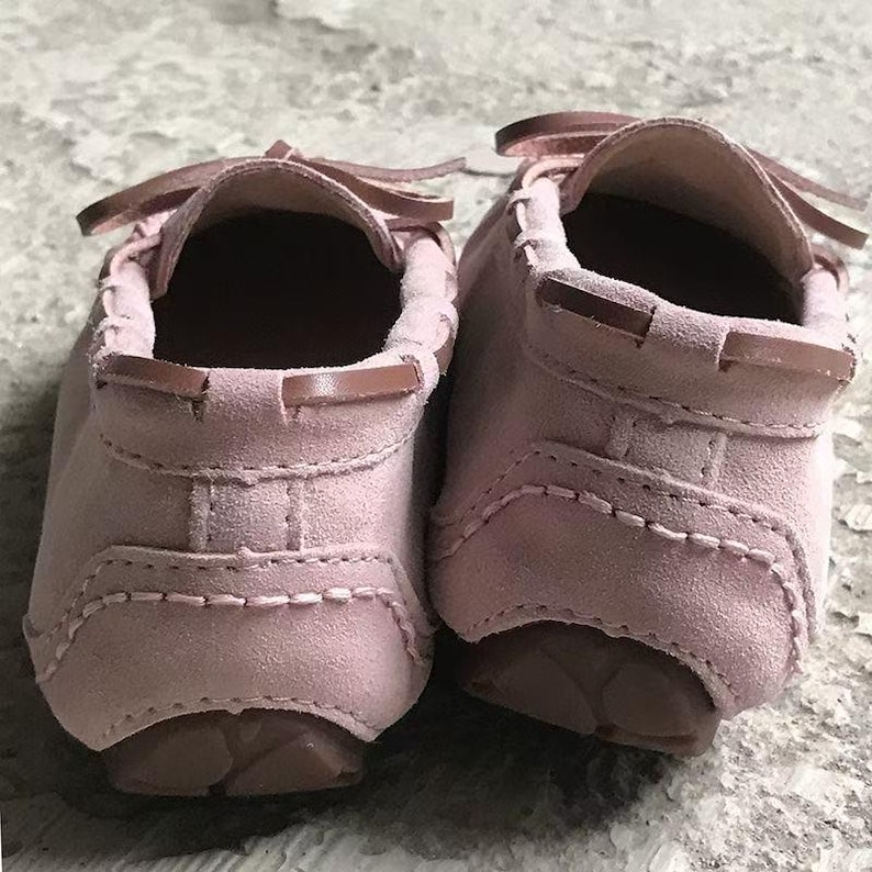 May include: A pair of pink suede moccasin-style shoes. The shoes have a rounded toe, a slip-on design, and a small bow detail at the top. The soles are a darker shade of pink. The shoes are designed for comfort and style.