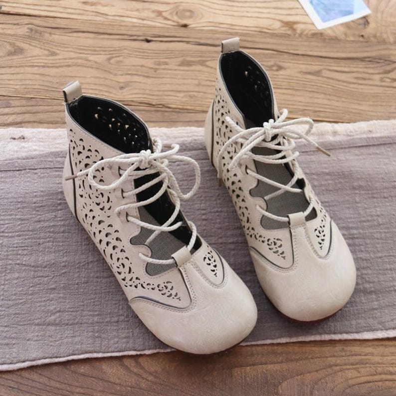 May include: A pair of cream-colored, lace-up ankle boots with intricate cut-out floral patterns. The boots have a rounded toe, a low heel, and white laces. The interior is black, and the boots are set on a gray surface.