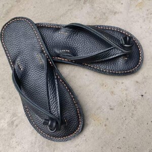 Puede incluir: Un par de chanclas de cuero negro con una superficie texturizada. Las sandalias tienen un dise&ntilde;o de correa de tanga y est&aacute;n cosidas con hilo marr&oacute;n claro alrededor de los bordes.
