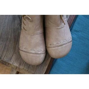 May include: Close-up of two taupe-colored leather boots. The boots have rounded toes, stitched detailing around the soles, and laces. The boots are on a wooden surface with a blue textile in the background. The boots appear to be new.