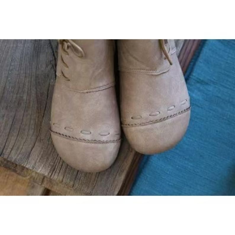May include: Close-up of two taupe-colored leather boots. The boots have rounded toes, stitched detailing around the soles, and laces. The boots are on a wooden surface with a blue textile in the background. The boots appear to be new.
