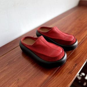 Puede incluir: Un par de zuecos de cuero rojo con suela negra. Los zuecos tienen una punta redondeada y un dise&ntilde;o sin cordones. Los zapatos est&aacute;n hechos de cuero texturizado y se apoyan sobre una superficie de madera.
