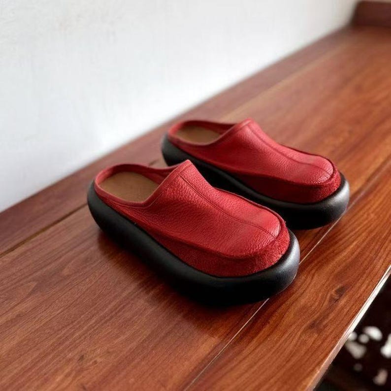 Puede incluir: Un par de zuecos de cuero rojo con suela negra. Los zuecos tienen una punta redondeada y un dise&ntilde;o sin cordones. Los zapatos est&aacute;n hechos de cuero texturizado y se apoyan sobre una superficie de madera.