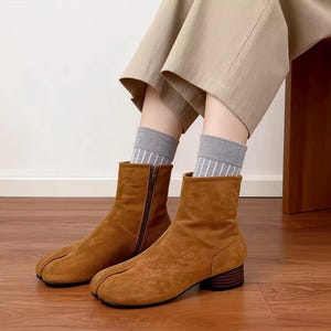 May include: Brown suede ankle boots with a split-toe design and a side zipper. The boots have a low, stacked heel and are worn with gray socks and tan pants. The boots are a warm, earthy tone, and the overall aesthetic is casual and stylish.