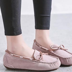 May include: A pair of dusty rose moccasin-style loafers with a bow detail on the vamp. The shoes have a stitched design around the toe and a brown trim. The shoes are worn with black pants.