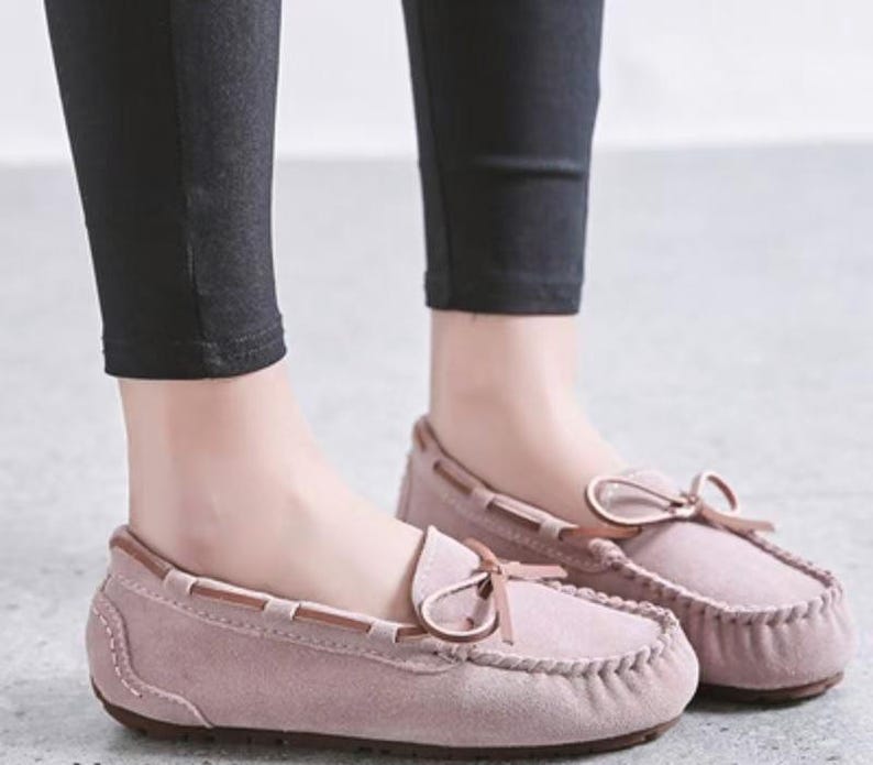 May include: A pair of dusty rose moccasin-style loafers with a bow detail on the vamp. The shoes have a stitched design around the toe and a brown trim. The shoes are worn with black pants.
