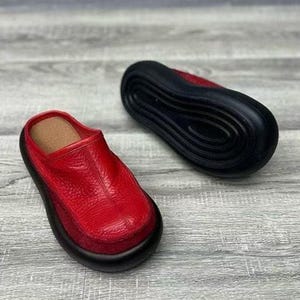 Puede incluir: Zuecos de cuero rojo con suela negra. Los zuecos tienen una parte superior de cuero texturizado y una plantilla marr&oacute;n. Un zueco est&aacute; en posici&oacute;n vertical y el otro est&aacute; boca abajo, mostrando la suela negra.
