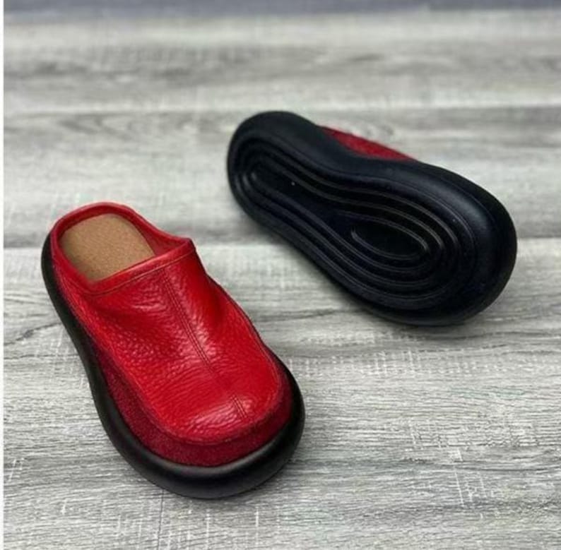 Puede incluir: Zuecos de cuero rojo con suela negra. Los zuecos tienen una parte superior de cuero texturizado y una plantilla marr&oacute;n. Un zueco est&aacute; en posici&oacute;n vertical y el otro est&aacute; boca abajo, mostrando la suela negra.
