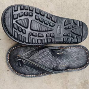 Puede incluir: Chanclas de cuero negro con suela texturizada y correa de dedo. La suela es negra con un dise&ntilde;o estampado. Las chanclas est&aacute;n cosidas con hilo marr&oacute;n claro alrededor de los bordes.