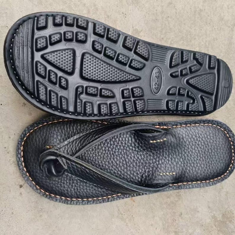 Puede incluir: Chanclas de cuero negro con suela texturizada y correa de dedo. La suela es negra con un dise&ntilde;o estampado. Las chanclas est&aacute;n cosidas con hilo marr&oacute;n claro alrededor de los bordes.