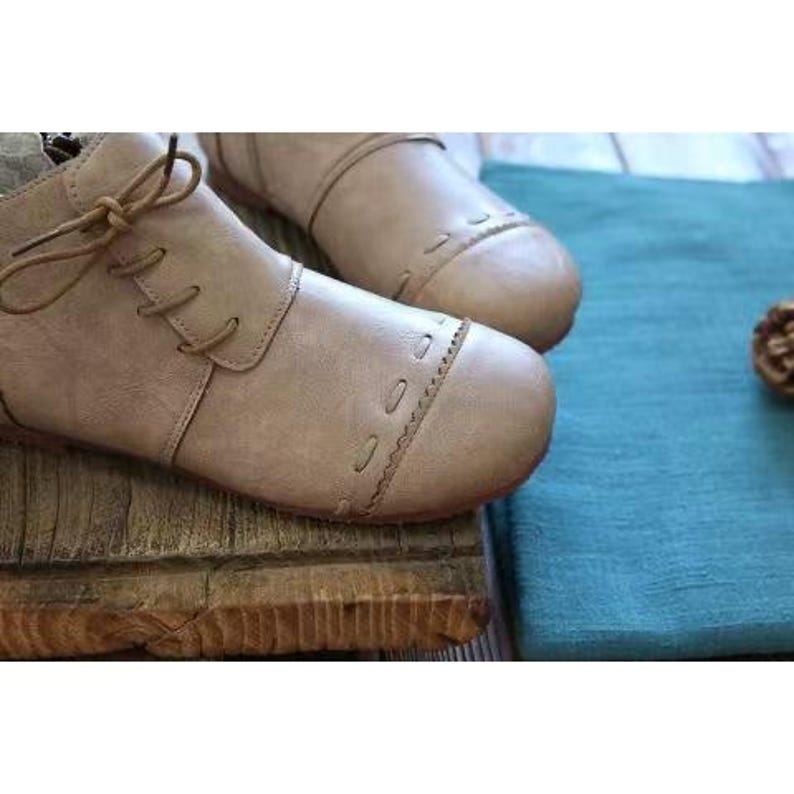May include: A pair of beige ankle boots with a lace-up design and decorative stitching on the toe. The boots are made of a soft, textured material and are placed on a wooden surface. The boots have a low heel and a round toe.