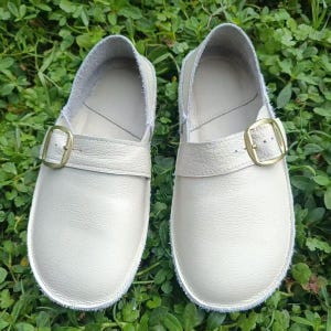 May include: A pair of off-white leather shoes with a buckle closure. The shoes have a rounded toe and a strap across the top with a gold-colored buckle. The shoes are set against a background of green grass.
