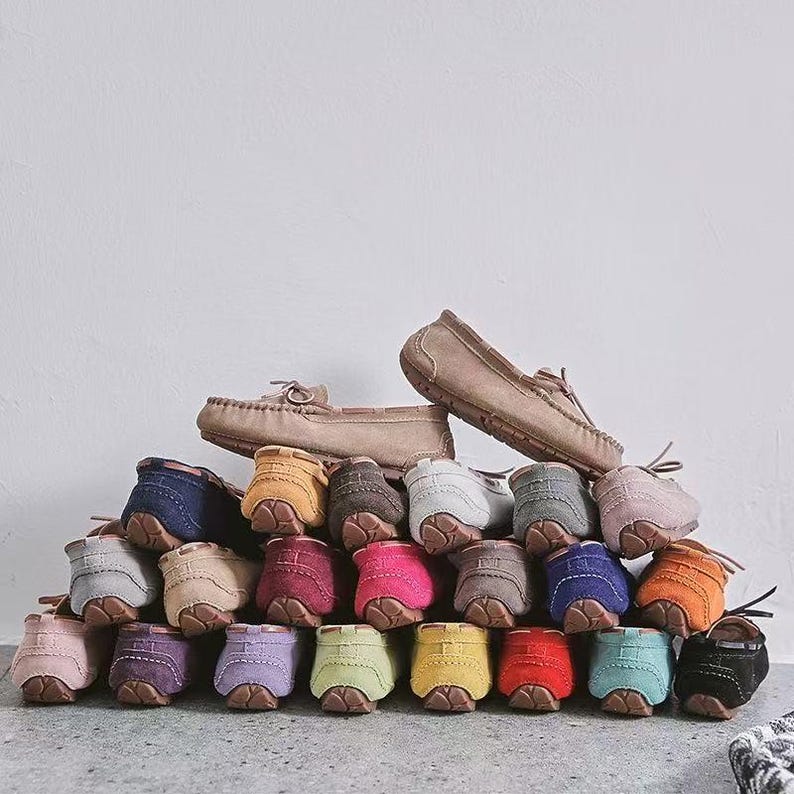 May include: A collection of colorful suede moccasin-style shoes in various shades, including pink, purple, yellow, and blue. Two pairs of tan-colored moccasins are at the top of the pile. The shoes are arranged in a pyramid shape.