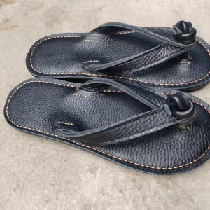 Puede incluir: Un par de chanclas de cuero negro con una superficie texturizada. Las sandalias tienen un dise&ntilde;o de correa de tanga y est&aacute;n cosidas con hilo marr&oacute;n claro alrededor de los bordes. Las correas presentan un detalle anudado.