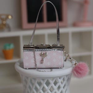 Puede incluir: Un bolso de mano rectangular en miniatura de color rosa claro con estampado de lunares blancos, con marco metálico plateado, asa plateada, correa de cadena plateada, un adorno de lazo plateado con una gema rosa y un pompón rosa esponjoso.