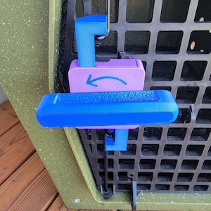 Puede incluir: Un mecanismo de cierre de plástico azul y morado fijado a un panel de rejilla negro. El componente morado tiene una flecha azul impresa que apunta a la izquierda. El mango azul es horizontal y está montado sobre una superficie verde oliva texturizada.