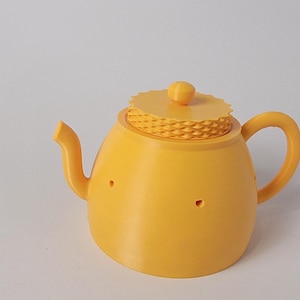 Puede incluir: Una tetera amarilla con tapa y asa a juego. La tetera tiene un cuerpo redondeado, un pico curvo y dos pequeños agujeros en la parte delantera. La tapa tiene un diseño texturizado y un pequeño pomo en la parte superior. La tetera es de material sólido.