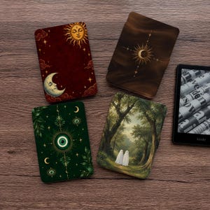 May include: Four rectangular e-reader cases featuring distinct designs: one deep red with a sun and moon, one dark brown with a celestial sun, one dark green with an eye motif, and one depicting two ghosts in a forest, alongside a black e-reader.