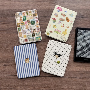 Puede incluir: Se muestran cuatro fundas para lectores electrónicos Kindle con diseños únicos sobre una superficie de madera. Las fundas presentan varios patrones, incluyendo un collage de arte enmarcado, arreglos florales, rayas azules con panqueques y un gato negro en una copa de martini.