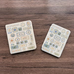 Può includere: Due fogli di adesivi rettangolari color bianco sporco che presentano una varietà di piccole etichette adesive individuali. Ogni etichetta mostra illustrazioni distinte di fiori, uccelli e motivi botanici in delicate tonalità di blu, verde, giallo e arancione.